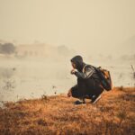 man in black jacket and black pants sitting on brown grass field near lake during daytime