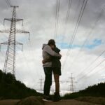 Couple hugging beneath power lines under a cloudy sky, showcasing a moment of affection and connection.