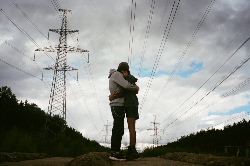 Couple hugging beneath power lines under a cloudy sky, showcasing a moment of affection and connection.