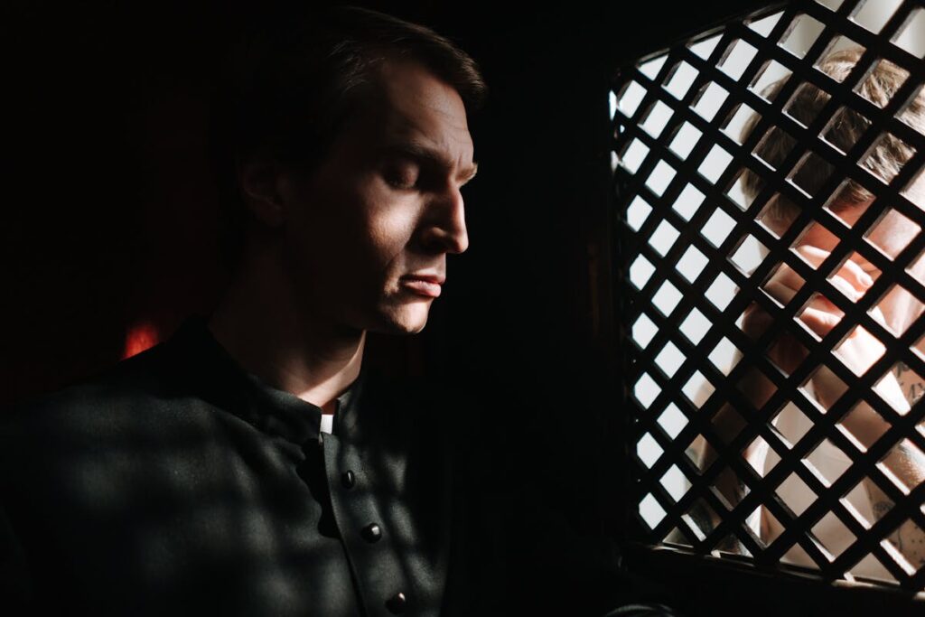 A priest listens to a penitent in a dimly lit confessional, embodying themes of faith and repentance.