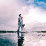A joyful couple embracing on the beach in wedding attire under a dramatic sky.