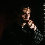 A priest pensively holds rosary beads inside a confessional, creating a spiritual ambiance with shadows.