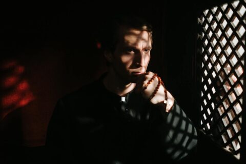 A priest pensively holds rosary beads inside a confessional, creating a spiritual ambiance with shadows.
