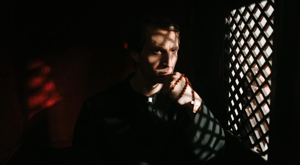 A priest pensively holds rosary beads inside a confessional, creating a spiritual ambiance with shadows.