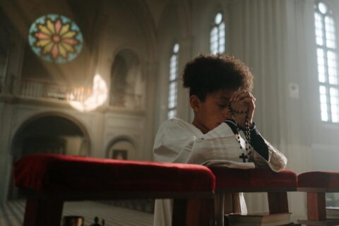 Child praying with rosary in a serene cathedral setting, sunlight through stained glass.