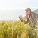 man reading book on beach near lake during daytime