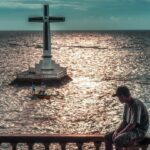 Man sits overlooking a large cross in the ocean.
