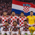 Hrvatska nogometna reprezentacija pozira na stadionu ispred velike hrvatske zastave dok navijači u pozadini pružaju podršku Vatrenima.