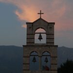 A tranquil view of a bell tower with a cross against sunset skies and distant mountains.