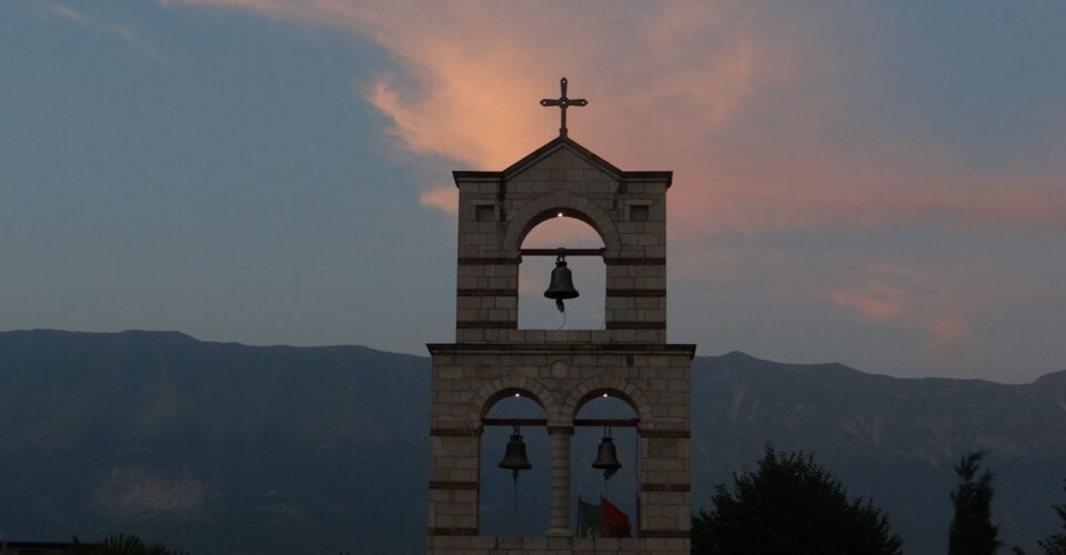 A tranquil view of a bell tower with a cross against sunset skies and distant mountains.
