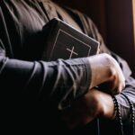 A priest holding a Bible by a sunlit window, symbolizing faith and spirituality.