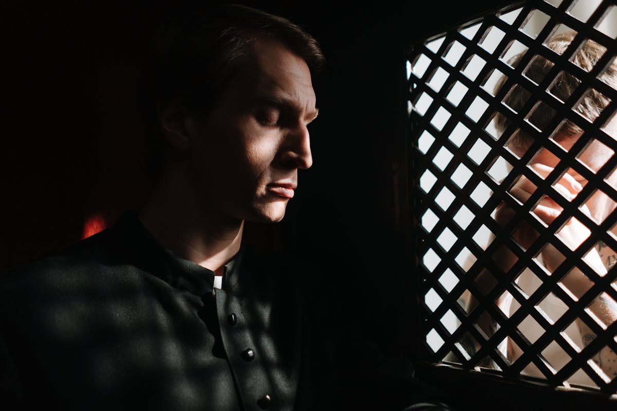 A priest listens to a penitent in a dimly lit confessional, embodying themes of faith and repentance.