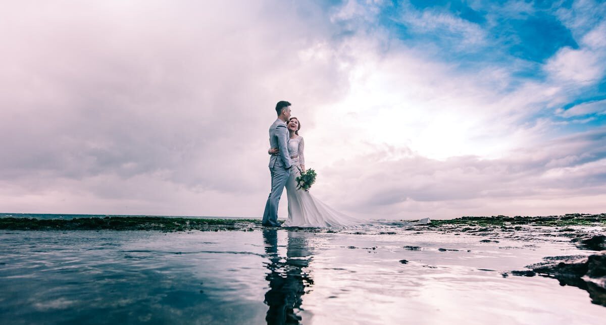A joyful couple embracing on the beach in wedding attire under a dramatic sky.
