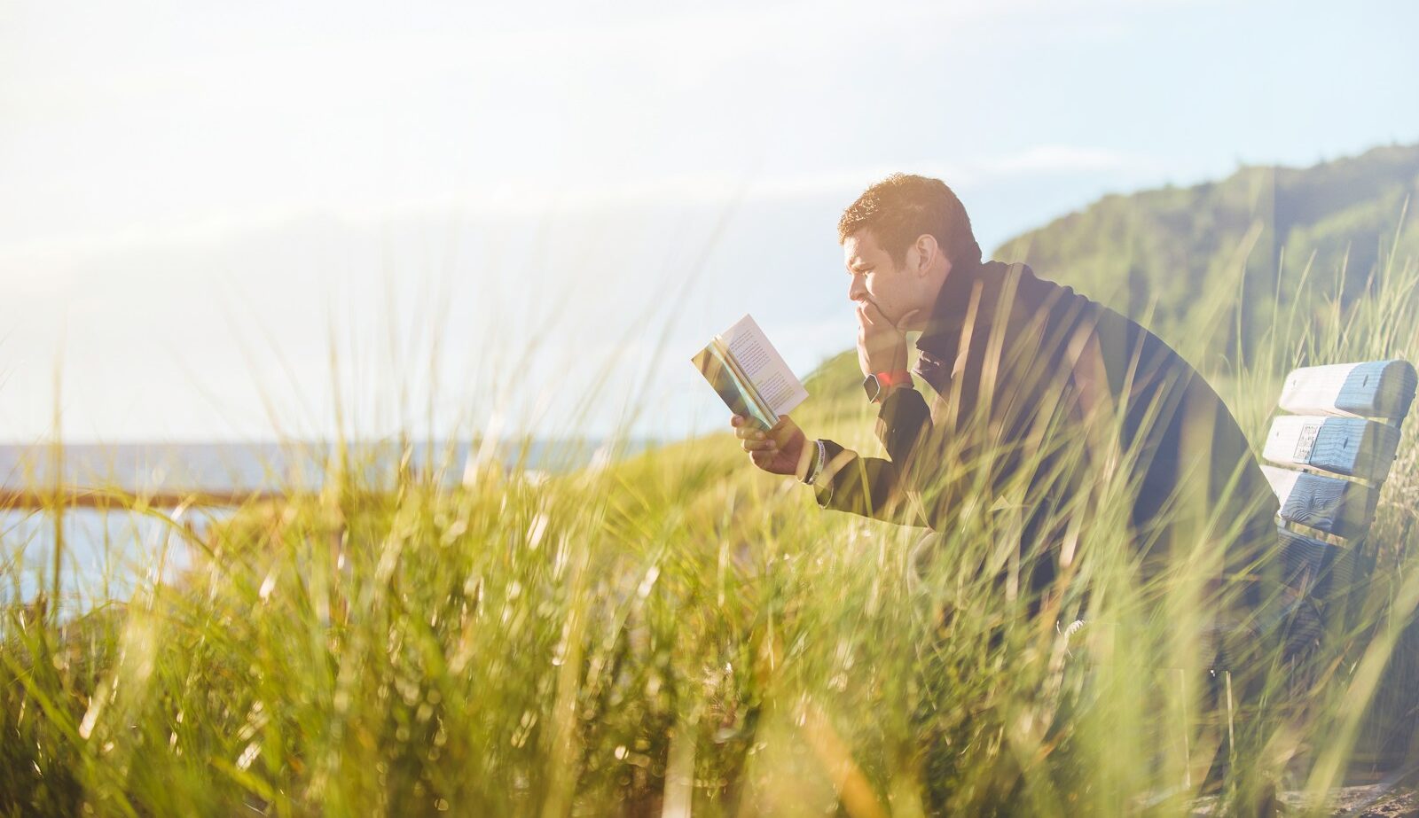 man reading book on beach near lake during daytime