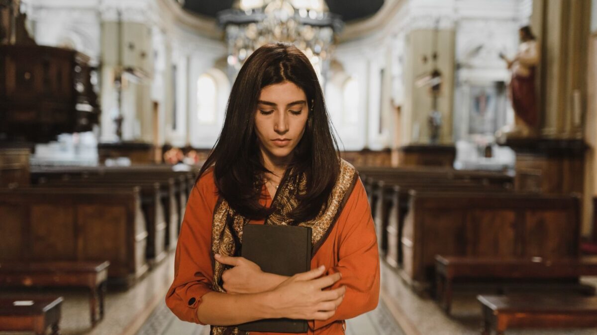 A woman in a church stands with a book, expressing contemplation and introspection.