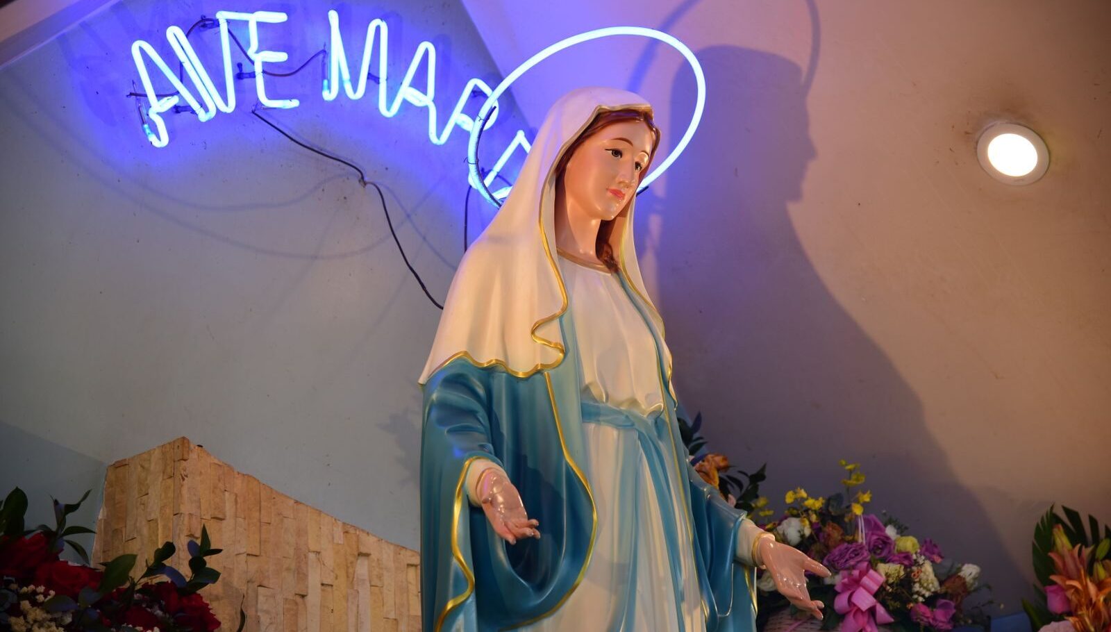 Elegant statue of Virgin Mary under a neon 'Ave Maria' sign with floral decor.