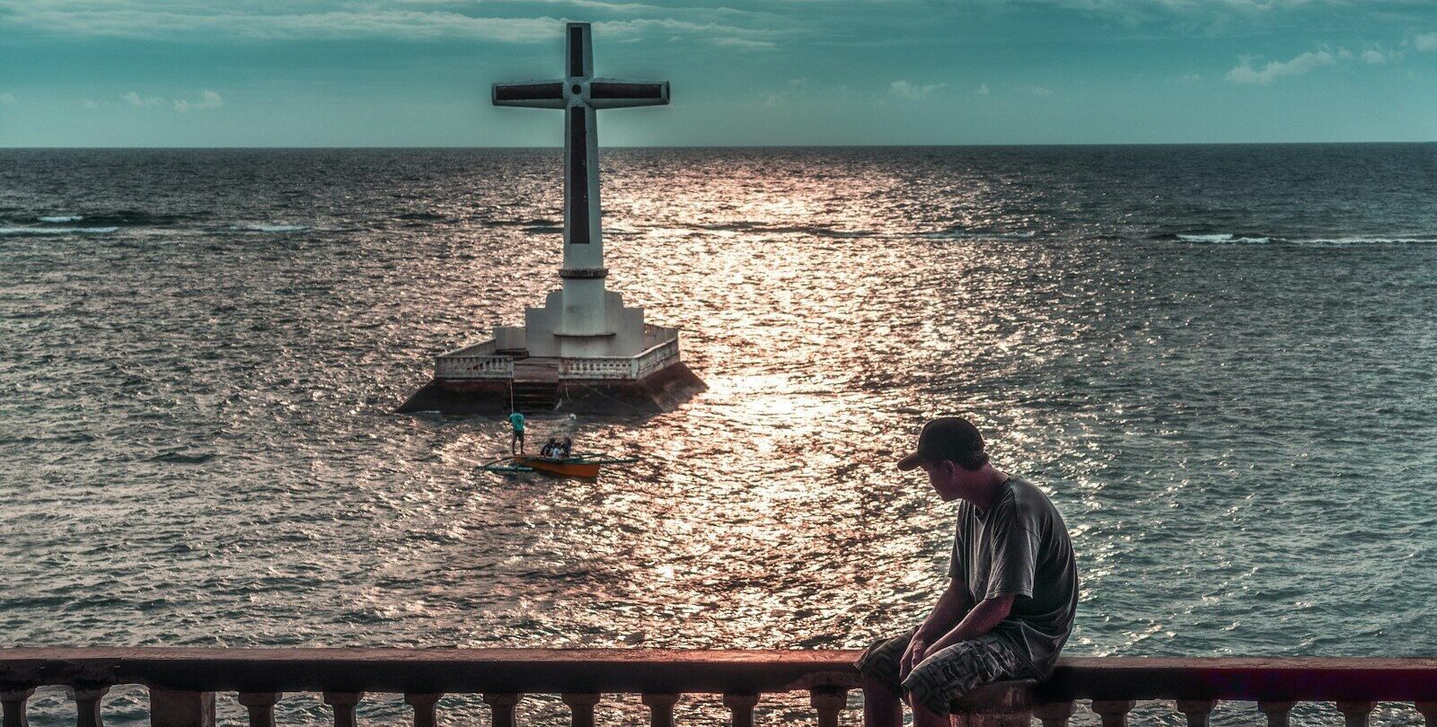 Man sits overlooking a large cross in the ocean.