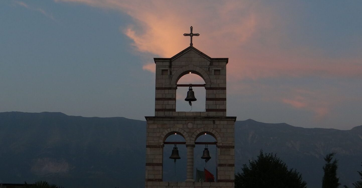 A tranquil view of a bell tower with a cross against sunset skies and distant mountains.