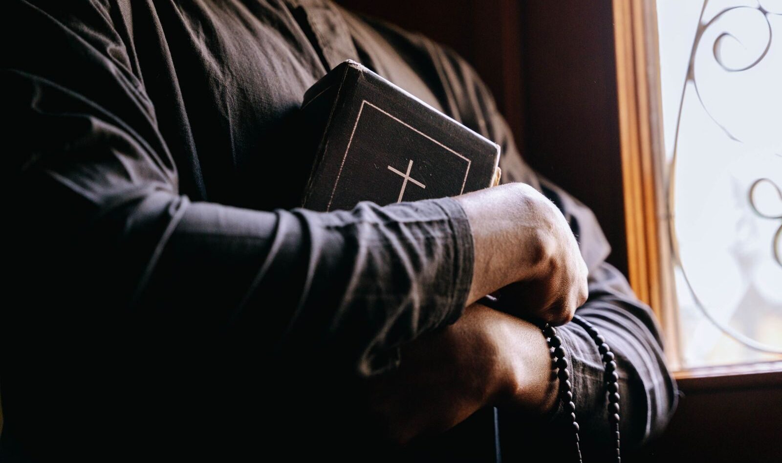 A priest holding a Bible by a sunlit window, symbolizing faith and spirituality.