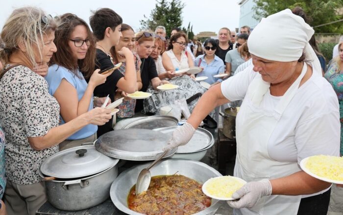 People enjoying a communal meal outdoors, served by a person in traditional attire.