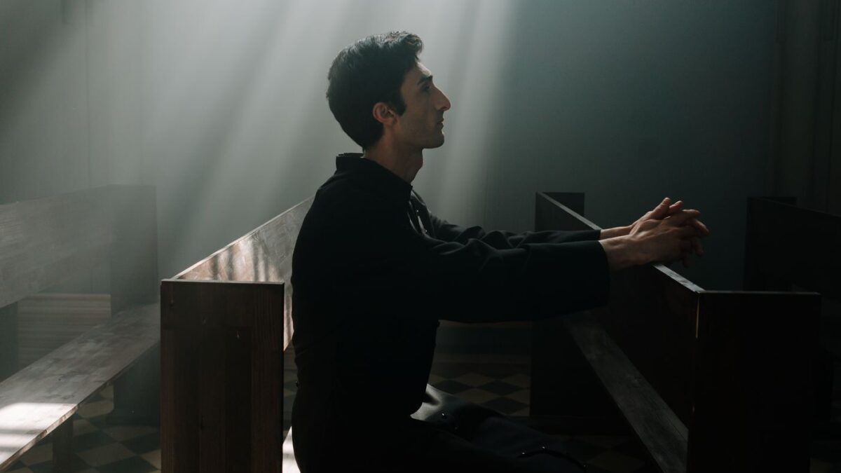 A man sits praying in sunlight inside a dramatic church setting.