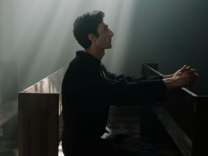 A man sits praying in sunlight inside a dramatic church setting.