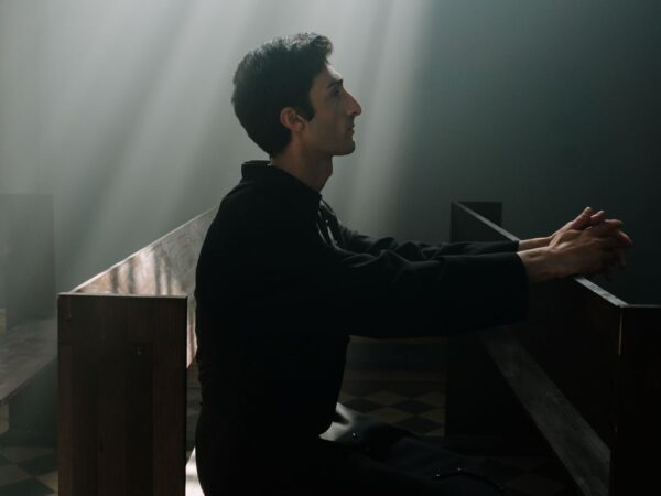A man sits praying in sunlight inside a dramatic church setting.