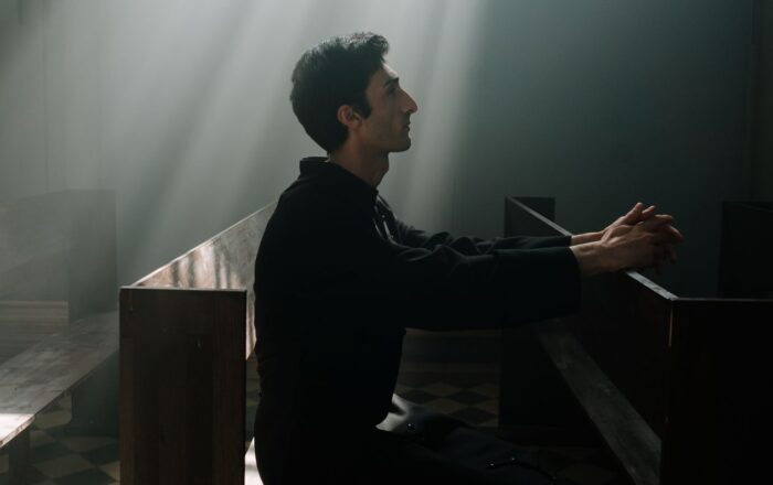 A man sits praying in sunlight inside a dramatic church setting.