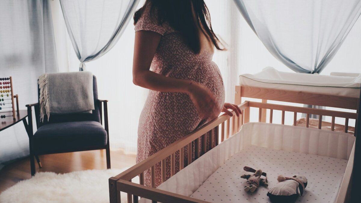 A pregnant woman standing by a wooden crib in a sunlit nursery